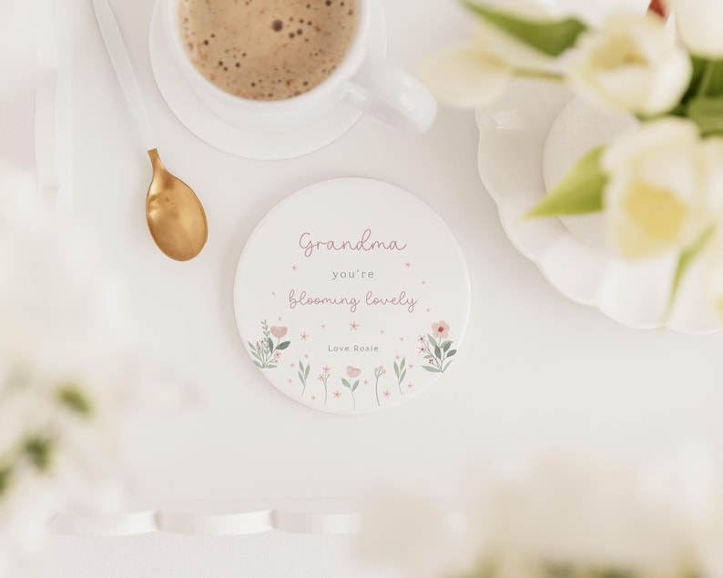 May include: A white ceramic coaster with the text "Grandma you're blooming lovely" in pink script, surrounded by floral illustrations. A gold spoon and a cup of coffee are visible in the background.
