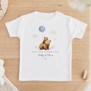 May include: White t-shirt featuring a watercolor illustration of two bears, one carrying the other, with a blue balloon. Text reads "Our First Father's Day" and "Daddy & Oliver" with a heart. A charming design for a child's tee.