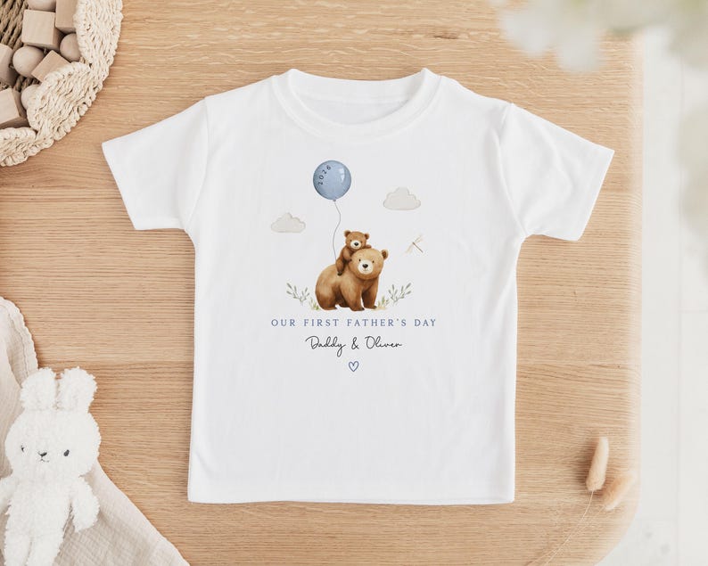 May include: White t-shirt featuring a watercolor illustration of two bears, one carrying the other, with a blue balloon. Text reads "Our First Father's Day" and "Daddy & Oliver" with a heart. A charming design for a child's tee.