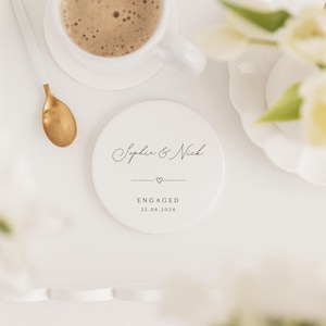 Puede incluir: Un posavasos de cer&aacute;mica blanca con los nombres "Sophie & Nick" en escritura elegante, un coraz&oacute;n y la palabra "ENGAGED" con la fecha 22.06.2026. Una taza de caf&eacute; y una cuchara dorada est&aacute;n cerca, con flores blancas en el fondo.