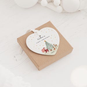 K&ouml;nnte beinhalten: Ein wei&szlig;es herzf&ouml;rmiges Keramikornament mit wei&szlig;em Band, das eine Illustration eines Weihnachtsbaums, eines Hirsches und eines roten Sacks zeigt. Der Text lautet Willow's FIRST CHRISTMAS und 2025, verziert mit kleinen goldenen Punkten.