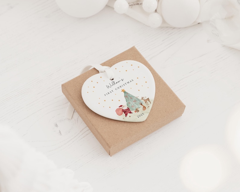 K&ouml;nnte beinhalten: Ein wei&szlig;es herzf&ouml;rmiges Keramikornament mit wei&szlig;em Band, das eine Illustration eines Weihnachtsbaums, eines Hirsches und eines roten Sacks zeigt. Der Text lautet Willow's FIRST CHRISTMAS und 2025, verziert mit kleinen goldenen Punkten.