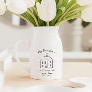 May include: A white ceramic pitcher with a handle, filled with white tulips. The pitcher is decorated with a black line drawing of a house and the words "My First Home", address, name, and date.