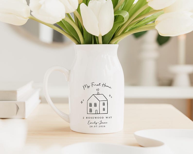 May include: A white ceramic pitcher with a handle, filled with white tulips. The pitcher is decorated with a black line drawing of a house and the words "My First Home", address, name, and date.