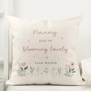 May include: Cream-colored square pillow with the text "Nanny you're blooming lovely" in pink script. Floral illustrations in pink and green are at the bottom. The text "Love Amelie" is also included.
