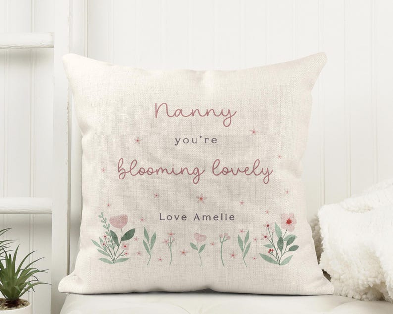 May include: Cream-colored square pillow with the text "Nanny you're blooming lovely" in pink script. Floral illustrations in pink and green are at the bottom. The text "Love Amelie" is also included.
