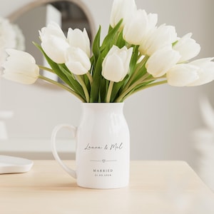 May include: A white ceramic pitcher with a handle, filled with white tulips. The pitcher has the names "Laura & Mel" and the word "MARRIED" with a date below. The flowers are fresh and vibrant, creating a bright and cheerful display.