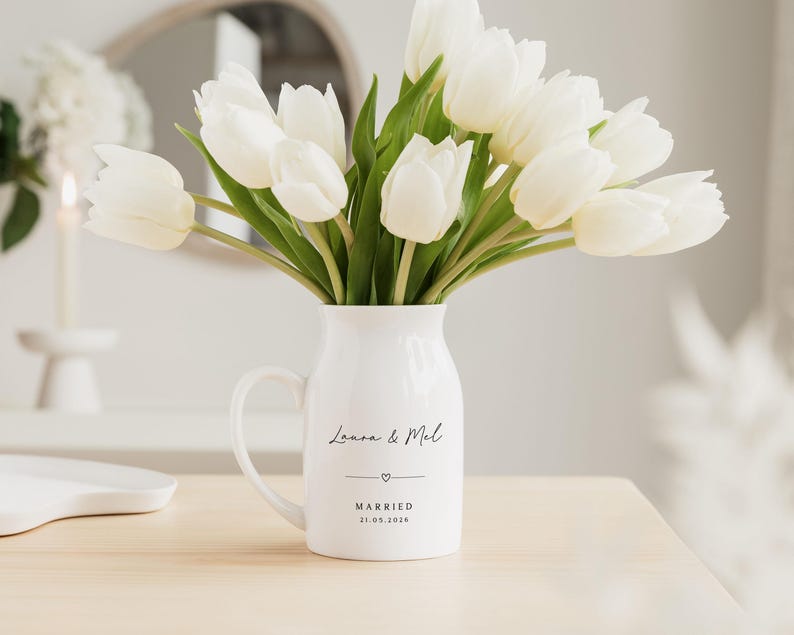 May include: A white ceramic pitcher with a handle, filled with white tulips. The pitcher has the names "Laura & Mel" and the word "MARRIED" with a date below. The flowers are fresh and vibrant, creating a bright and cheerful display.