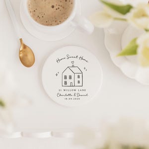 May include: A white ceramic coaster with a black line drawing of a house, the words "Home Sweet Home", and the address "21 Willow Lane". A cup of coffee, a gold spoon, and flowers are also in the image.