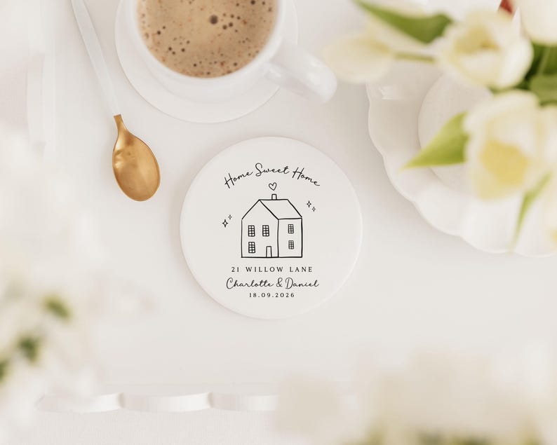 May include: A white ceramic coaster with a black line drawing of a house, the words "Home Sweet Home", and the address "21 Willow Lane". A cup of coffee, a gold spoon, and flowers are also in the image.