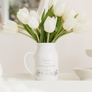 May include: A white ceramic pitcher with a handle, filled with white tulips. The pitcher has the text "Nana you're blooming lovely" with floral illustrations. A white candle and small bowl are also visible.