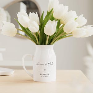 Puede incluir: Una jarra de cer&aacute;mica blanca con asa, llena de tulipanes blancos. La jarra tiene las palabras "Laura & Mel" y "ENGAGED 21.05.2026" impresas. Las flores frescas crean una exhibici&oacute;n brillante.