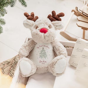 May include: A plush reindeer toy with brown antlers, a red nose, and cream-colored fur. The reindeer is sitting in a wooden crate. The text "My First Christmas" and a Christmas tree design are printed on the reindeer's chest, along with the name "Emilia" and the year "2025".