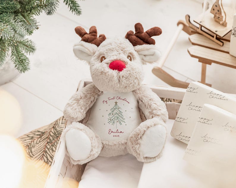 May include: A plush reindeer toy with brown antlers, a red nose, and cream-colored fur. The reindeer is sitting in a wooden crate. The text "My First Christmas" and a Christmas tree design are printed on the reindeer's chest, along with the name "Emilia" and the year "2025".
