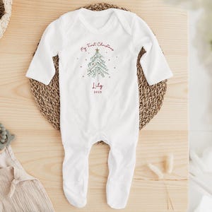 May include: White baby onesie with long sleeves and attached feet. The onesie features a Christmas tree design with the text "My First Christmas" and the name "Lily" with the year "2025" in red.