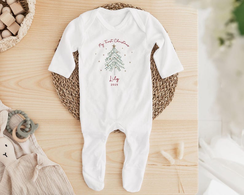 May include: White baby onesie with long sleeves and attached feet. The onesie features a Christmas tree design with the text "My First Christmas" and the name "Lily" with the year "2025" in red.
