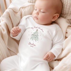 May include: A sleeping baby wearing a white onesie with long sleeves. The onesie has a Christmas tree design and the text "My First Christmas" and "Emilia 2025" in burgundy. The baby is in a wooden crib with a beige blanket.