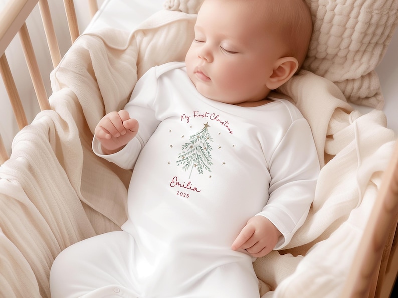 May include: A sleeping baby wearing a white onesie with long sleeves. The onesie has a Christmas tree design and the text "My First Christmas" and "Emilia 2025" in burgundy. The baby is in a wooden crib with a beige blanket.