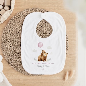May include: White baby bib with a watercolor illustration of two brown bears, one holding a pink balloon. The text reads "OUR FIRST FATHER'S DAY" and "Daddy & Olivia". The bib is on a woven mat.