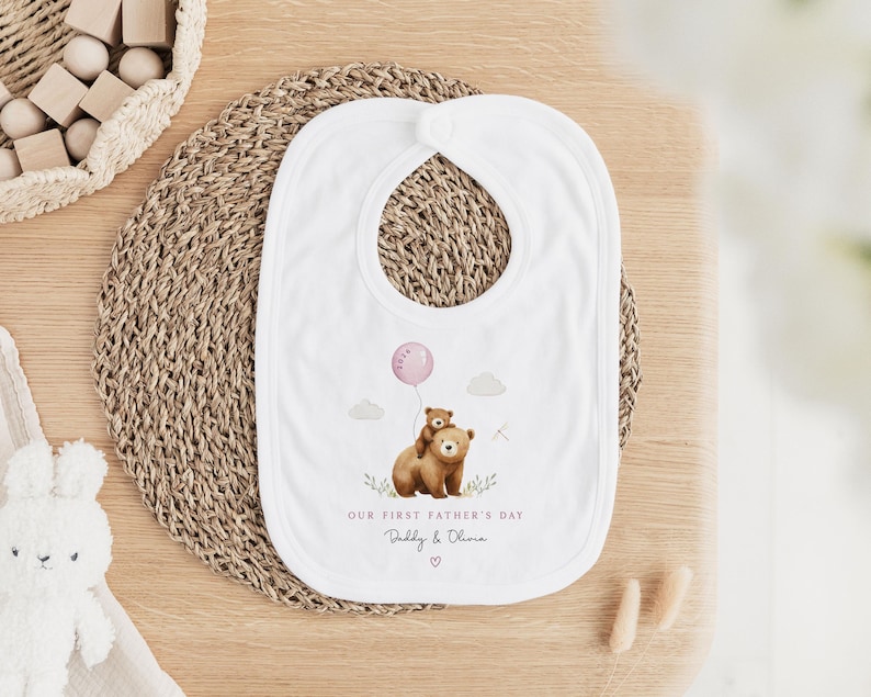 May include: White baby bib with a watercolor illustration of two brown bears, one holding a pink balloon. The text reads "OUR FIRST FATHER'S DAY" and "Daddy & Olivia". The bib is on a woven mat.