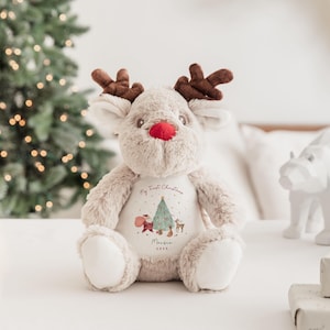 K&ouml;nnte beinhalten: Ein beiges Pl&uuml;sch-Rentier mit dunkelbraunem Geweih und einer leuchtend roten Nase. Das Rentier hat einen wei&szlig;en Brustbereich, der mit einem Weihnachtsbaum, dem Weihnachtsmann, einem kleinen Reh und dem Text My First Christmas, Maisie, 2023 bedruckt ist.