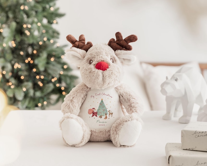 K&ouml;nnte beinhalten: Ein beiges Pl&uuml;sch-Rentier mit dunkelbraunem Geweih und einer leuchtend roten Nase. Das Rentier hat einen wei&szlig;en Brustbereich, der mit einem Weihnachtsbaum, dem Weihnachtsmann, einem kleinen Reh und dem Text My First Christmas, Maisie, 2023 bedruckt ist.