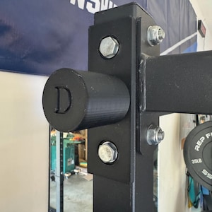Puede incluir: Primer plano de un rack de halterofilia negro con pernos y una tapa de extremo cilíndrica con la letra "D". El rack está hecho de metal negro y está atornillado. Una pancarta con el texto "MEN'S" es visible en el fondo.