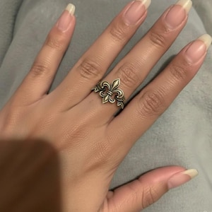 May include: A bronze-colored ring featuring a fleur-de-lis design. The ring is worn on a finger, showcasing the intricate details of the floral emblem. The ring's design includes stylized leaves, adding to its decorative appeal.