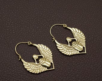 May include: A pair of gold-colored hoop earrings. Each earring features a winged scarab beetle design. The wings are detailed with feather patterns, and the beetle is centered within a square frame. The earrings are displayed on a brown surface.