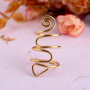 May include: A gold-colored, wire-wrapped ring with a unique design. The ring features a spiral at the top, a wavy middle section, and a triangular shape at the bottom. The ring is likely made of metal and is designed to be worn on a finger.