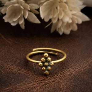 May include: A gold-colored adjustable ring with a cluster of small, round beads at the center. The ring is set against a dark brown, textured surface, with white flowers in the background. The ring's design is simple yet elegant.
