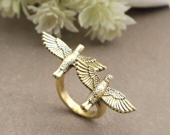 May include: A gold-coloured ring featuring two detailed bird figures with outstretched wings. The birds are positioned to appear as if they are in flight, creating a dynamic design. The ring is set against a neutral background.