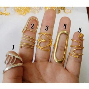 Op de afbeelding: Een hand toont vijf ringen in verschillende ontwerpen. De ringen zijn goud- en zilverkleurig. De ringen omvatten een zilveren golfontwerp, een gouden spiraal, een gouden hart, een gouden ovaal en een gouden spiraalontwerp.