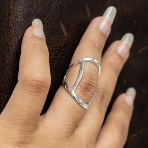 May include: A silver-colored ring with a geometric design. The ring features a triangular shape with a vertical and diagonal bar. The ring is worn on a finger against a brown background.