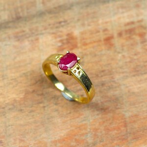 May include: A gold ring featuring a vibrant oval-cut ruby gemstone. The ring's band is a polished gold, with decorative cutouts near the ruby setting. The ruby is a deep red color, set in a classic prong setting.