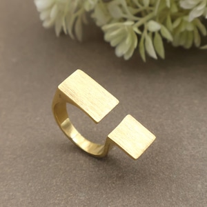 May include: A gold ring with a contemporary design. The ring has two unconnected rectangular features, creating an open design. The ring is made of a brushed metal material.