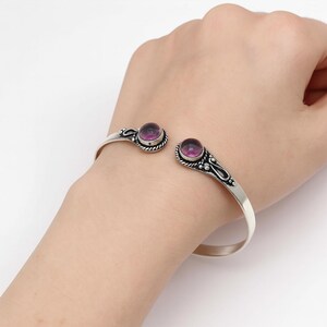 Peut inclure: Un bracelet manchette en argent avec deux pierres pr&eacute;cieuses rondes et violettes. Le bracelet pr&eacute;sente des d&eacute;tails d&eacute;coratifs autour des pierres pr&eacute;cieuses et sur les bords. La conception ouverte permet de l'ajuster &agrave; diff&eacute;rentes tailles de poignet.
