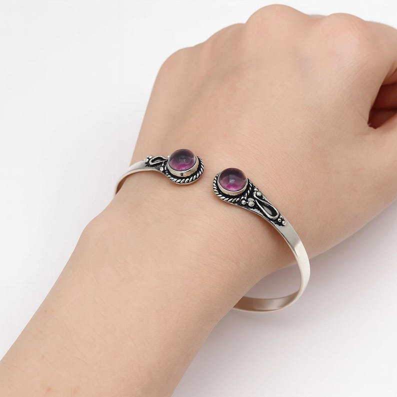 Peut inclure: Un bracelet manchette en argent avec deux pierres pr&eacute;cieuses rondes et violettes. Le bracelet pr&eacute;sente des d&eacute;tails d&eacute;coratifs autour des pierres pr&eacute;cieuses et sur les bords. La conception ouverte permet de l'ajuster &agrave; diff&eacute;rentes tailles de poignet.