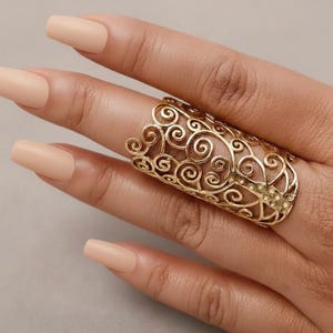 May include: A gold-colored filigree ring with an ornate, swirling design. The ring is worn on a finger with light-toned, polished nails. The ring's intricate pattern and warm color create a striking contrast against the skin.