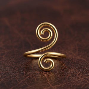 May include: A gold-colored ring with a spiral design. The ring features a double spiral, with one spiral at the top and another at the base, connected by a curved band. The ring is made of a shiny, metallic material.