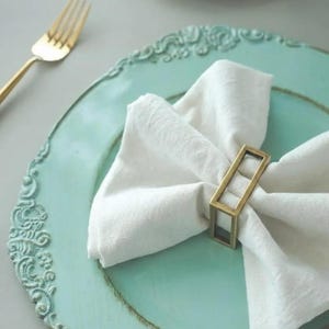 May include: A white linen napkin is folded and held by a gold-colored rectangular napkin ring on a turquoise plate with ornate edges. A gold fork rests on the left side of the plate.