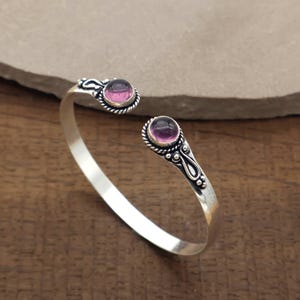 Peut inclure: Un bracelet manchette argent&eacute; avec deux pierres pr&eacute;cieuses rondes, serties &agrave; griffes, de couleur rose-violet. Le bracelet pr&eacute;sente des d&eacute;tails d&eacute;coratifs autour des pierres et le long des extr&eacute;mit&eacute;s ouvertes. Les pierres mesurent environ 0,8 cm de diam&egrave;tre.