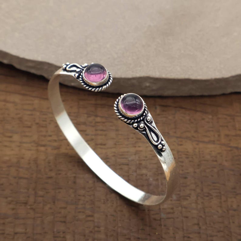 Peut inclure: Un bracelet manchette argent&eacute; avec deux pierres pr&eacute;cieuses rondes, serties &agrave; griffes, de couleur rose-violet. Le bracelet pr&eacute;sente des d&eacute;tails d&eacute;coratifs autour des pierres et le long des extr&eacute;mit&eacute;s ouvertes. Les pierres mesurent environ 0,8 cm de diam&egrave;tre.