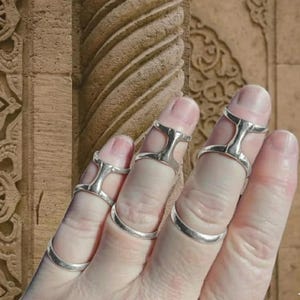 May include: Silver finger rings with a unique design. The rings have a band that goes around the base of the finger and a decorative element that extends up the finger. The rings are worn on multiple fingers.