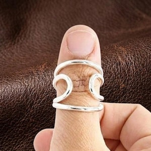 Puede incluir: Un anillo de plata con un diseño ancho y abierto que presenta dos bandas horizontales conectadas por dos elementos verticales curvos en forma de onda. El metal tiene un acabado pulido y reflectante, creando una estructura continua.