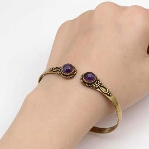Peut inclure: Un bracelet manchette dor&eacute; avec deux pierres pr&eacute;cieuses rondes, violettes fonc&eacute;es. Le bracelet est de conception ouverte et pr&eacute;sente des d&eacute;tails d&eacute;coratifs autour des pierres. Le bracelet est port&eacute; sur un poignet.