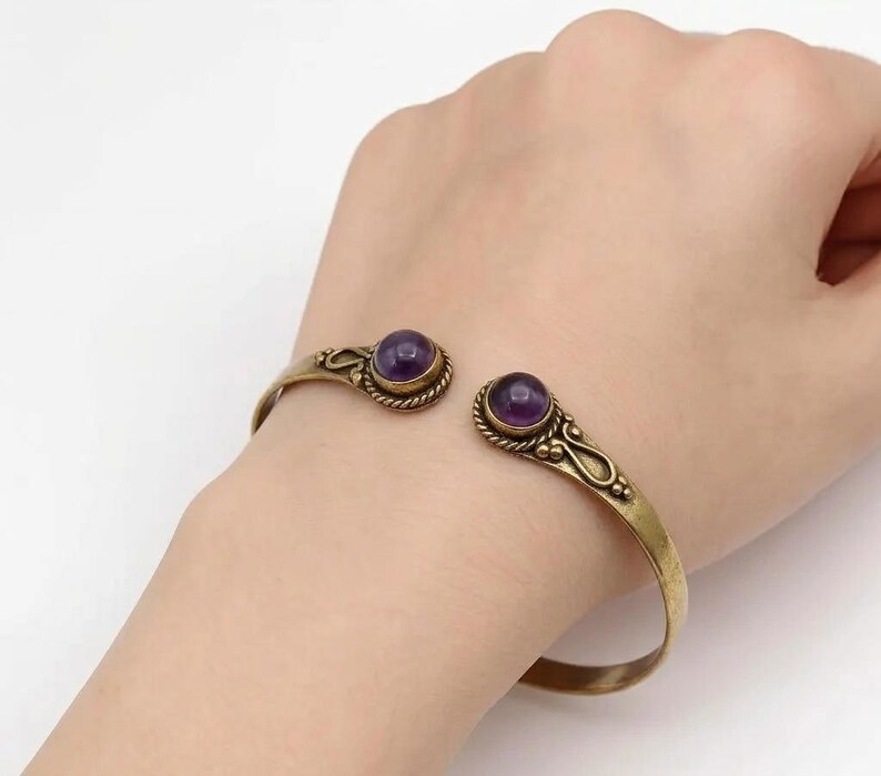 Peut inclure: Un bracelet manchette dor&eacute; avec deux pierres pr&eacute;cieuses rondes, violettes fonc&eacute;es. Le bracelet est de conception ouverte et pr&eacute;sente des d&eacute;tails d&eacute;coratifs autour des pierres. Le bracelet est port&eacute; sur un poignet.