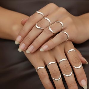May include: A selection of silver rings in a range of curved shapes. The rings are displayed on several fingers, highlighting a minimalist and contemporary jewellery style. The rings have a polished finish.
