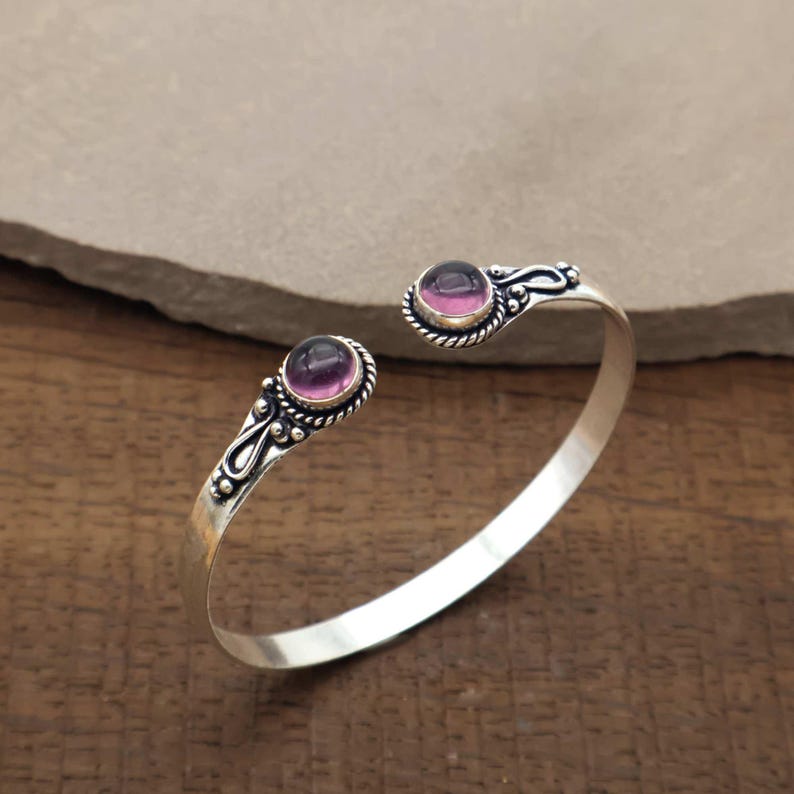 Peut inclure: Un bracelet manchette argent&eacute; avec deux pierres pr&eacute;cieuses rondes et violettes. Les pierres sont serties dans une bordure d&eacute;corative en forme de corde. Le bracelet est ouvert, avec les pierres &agrave; chaque extr&eacute;mit&eacute;. Sur une surface en bois.