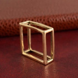 May include: A gold-colored, rectangular ring with an open, geometric design. The ring is made of a shiny metal and has a hollow, box-like shape. The ring is shown on a red surface.